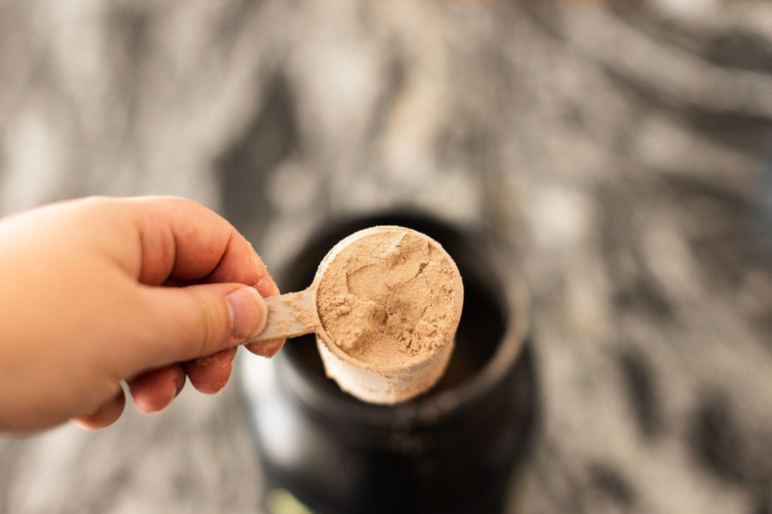 Person holding chocolate whey protein powder in a measuring scoop above a kitchen counter